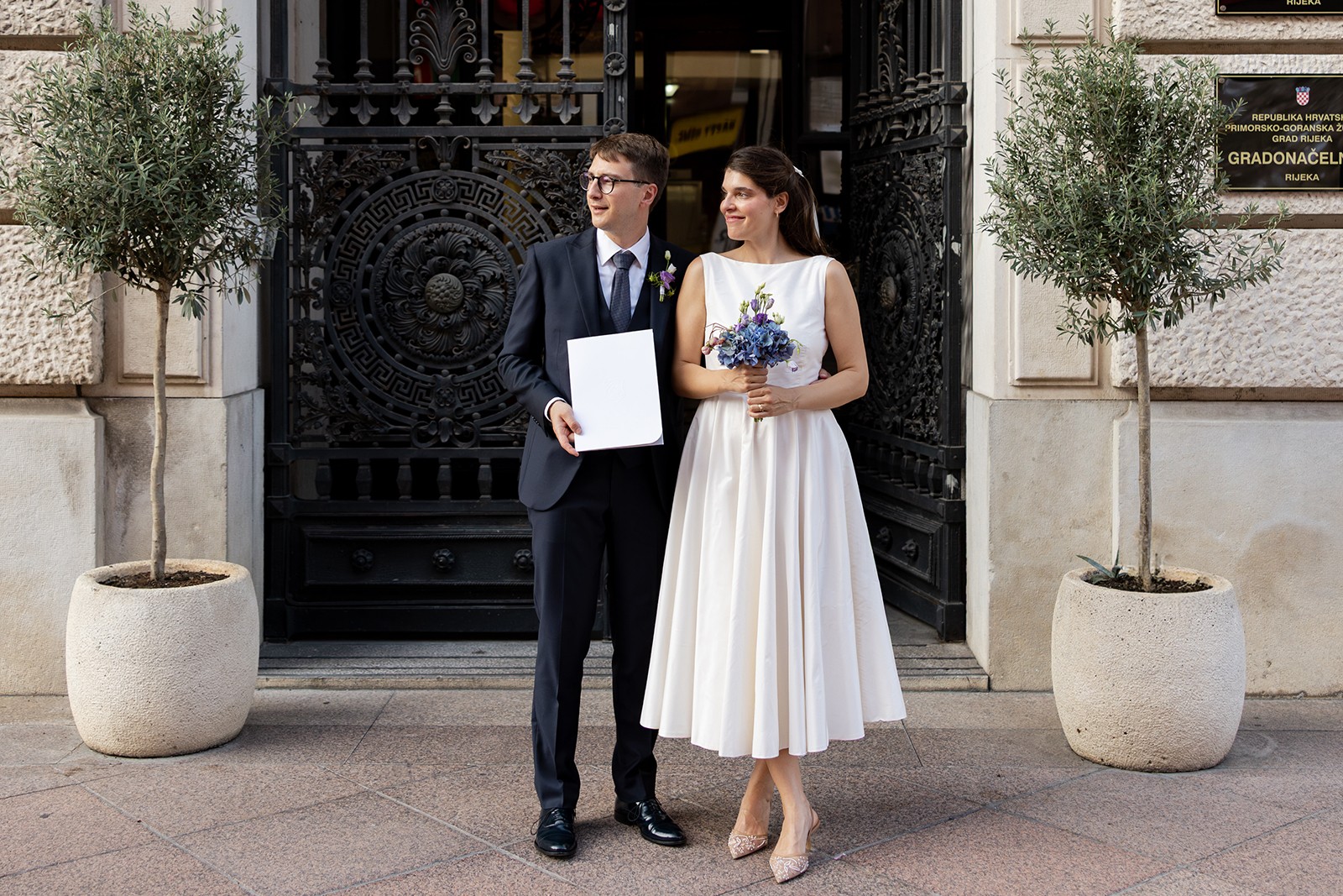 Bride and groom holding marriage certificate infront of a stone building that has a large black gate and two olive trees