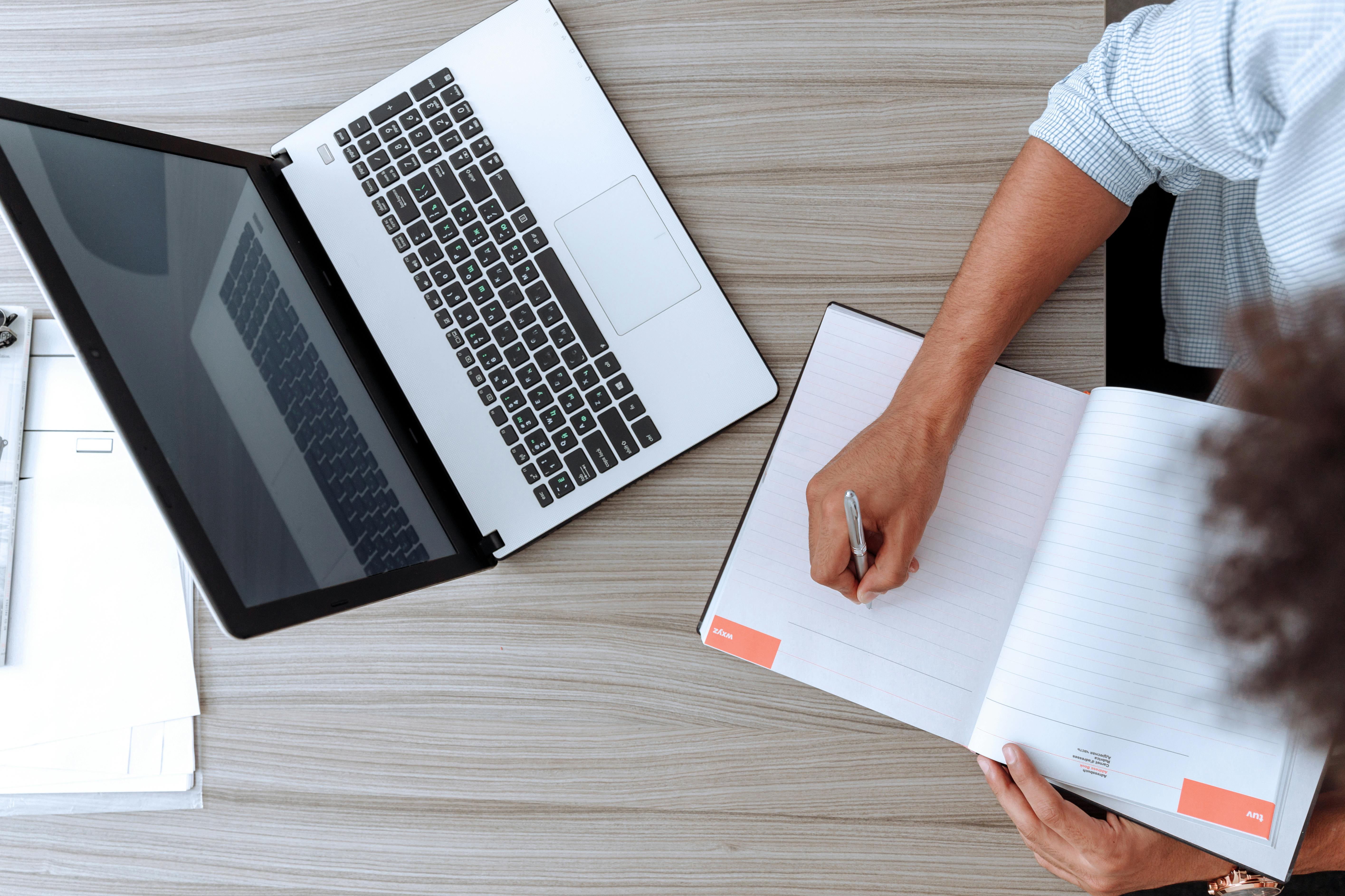 woman writing something in notebook with open laptop on desk