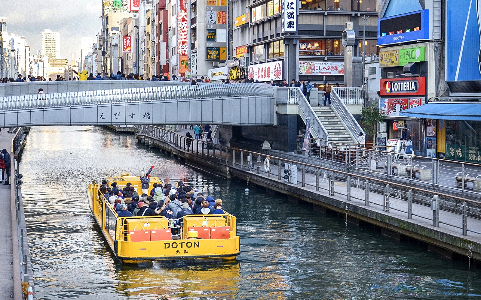 ਓਸਾਕਾ, ਜਪਾਨ ਵਿੱਚ ਡੋਟਨਬੋਰੀ ਪੁਲ ਦੇ ਨੀچے ਟੋਮਬੋਰੀ ਨਦੀ ਦੀ ਕ੍ਰੂਜ਼ ਬੋਟ ਗੁਜ਼ਰ ਰਹੀ ਹੈ।