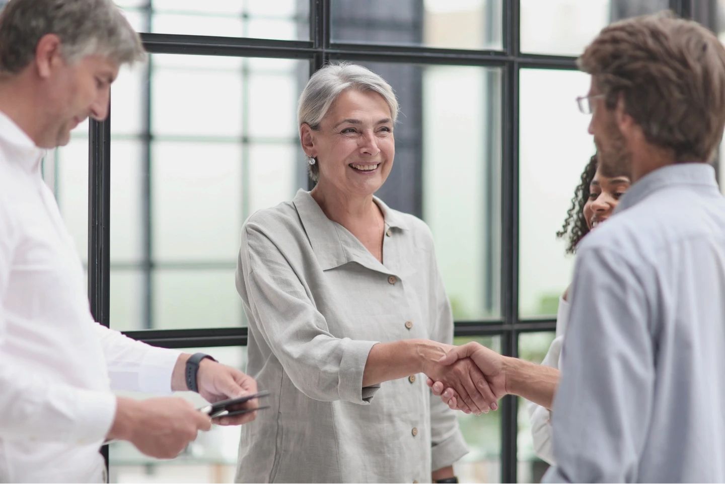 A diverse group of professionals engages in a friendly handshake and conversation in a modern office with large, sunlit windows, conveying a sense of teamwork and collaboration.