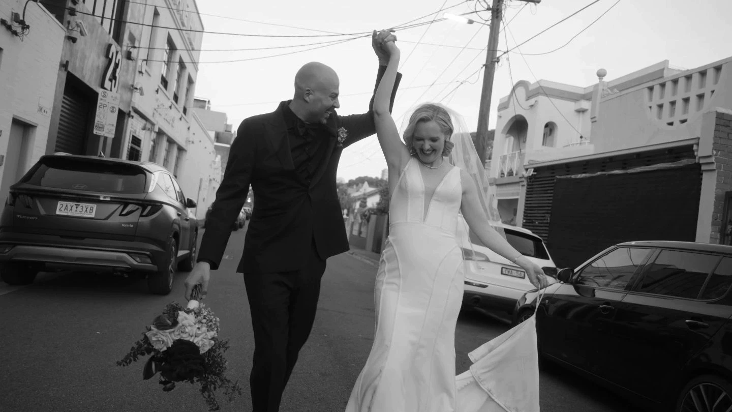 A joyful couple, dressed in a formal suit and elegant wedding gown, celebrate their wedding, holding hands and smiling while walking down an urban street adorned with historic architecture and vintage cars.