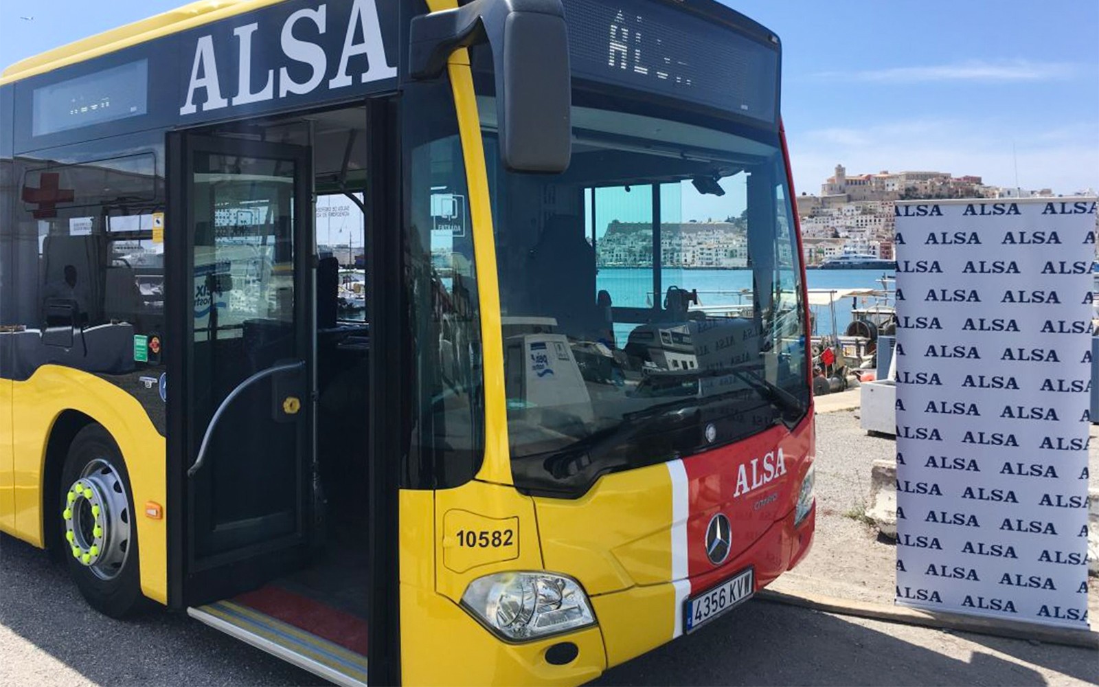 Bus ALSA kuning di pelabuhan Ibiza dengan latar belakang pemandangan kota.