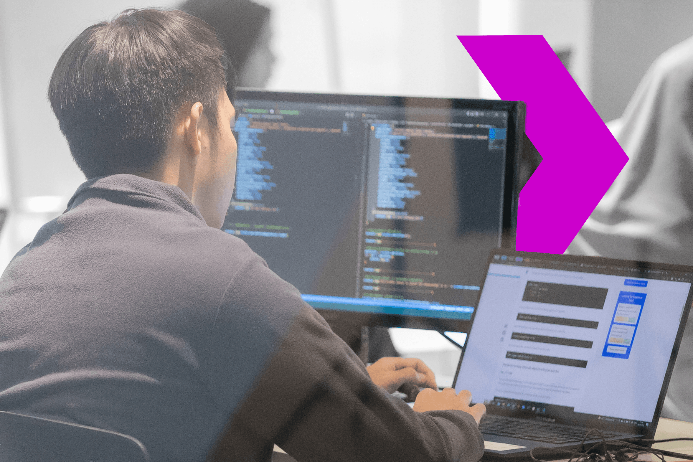 A programmer viewing code on dual monitors and typing on a laptop, focused on software development tasks in a modern workspace.