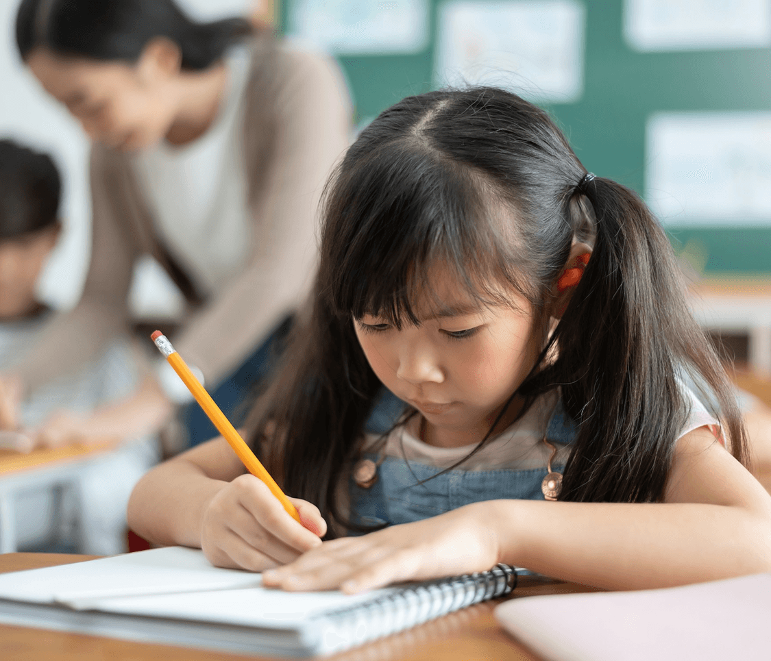 A child focused on writing in a notebook, with a teacher assisting in the background. Classroom setting.