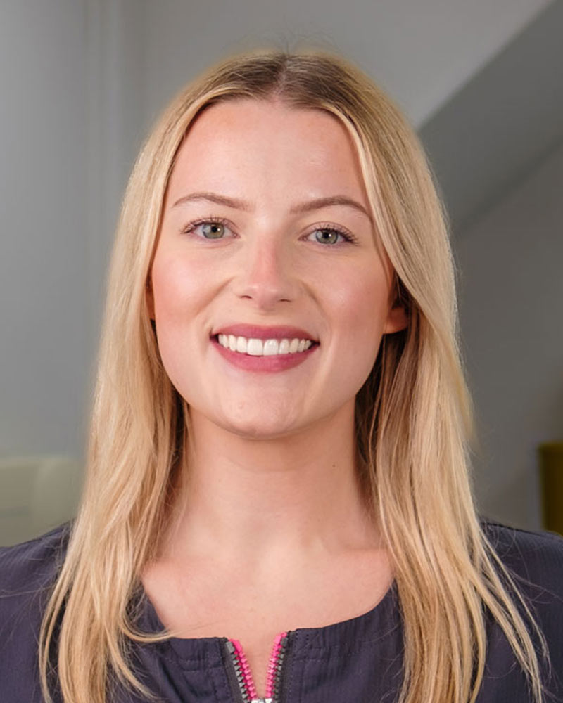 A portrait of Flora, an Associate Dentist at Cricklade Dental Practice, smiling and wearing a dark grey scrub top with yellow embroidered text that reads "Flora" on the left and "Cricklade Dental Practice" on the right. She is standing in a dental surgery with equipment visible in the background.