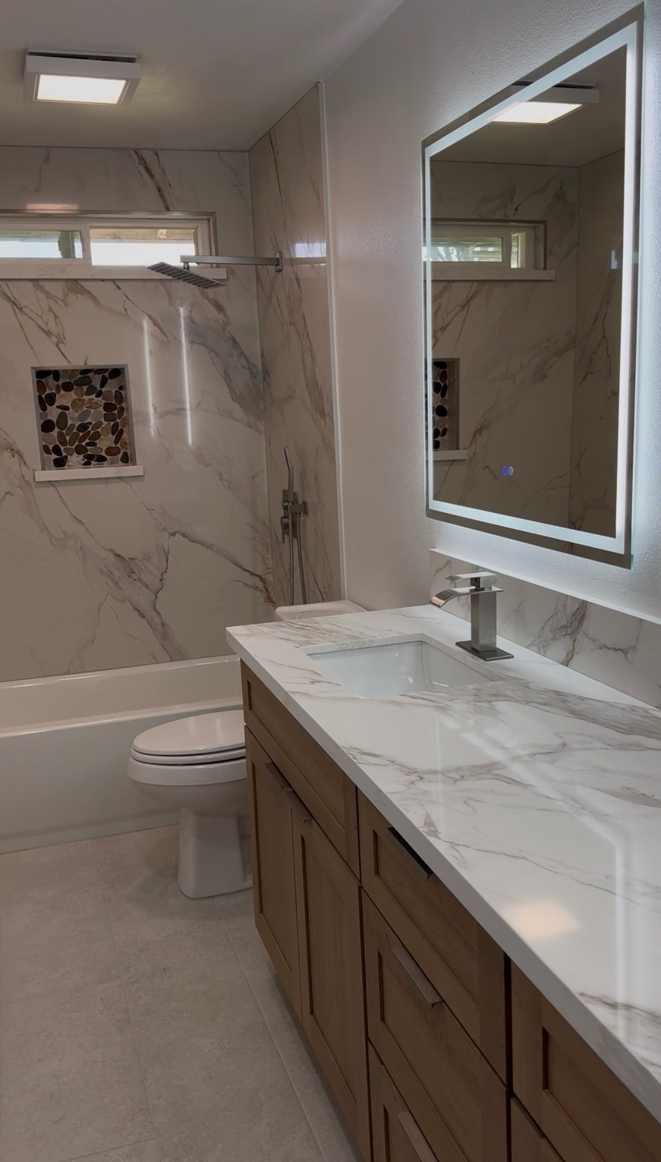 Finished bathroom with marble-look tile, a wood vanity, and a lighted mirror by All American Renovations