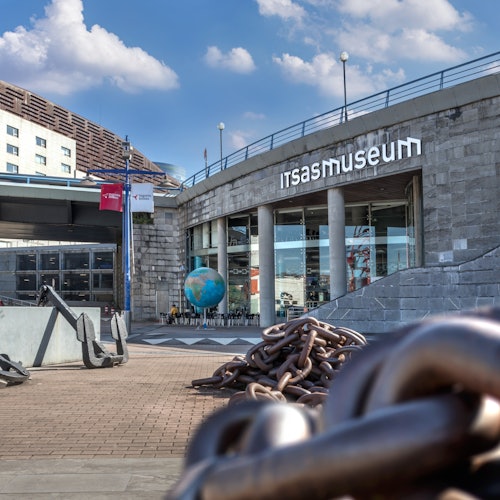 Entrance of Itsasmuseum with large anchors, heavy chains, flags, and a globe sculpture outside the building.
