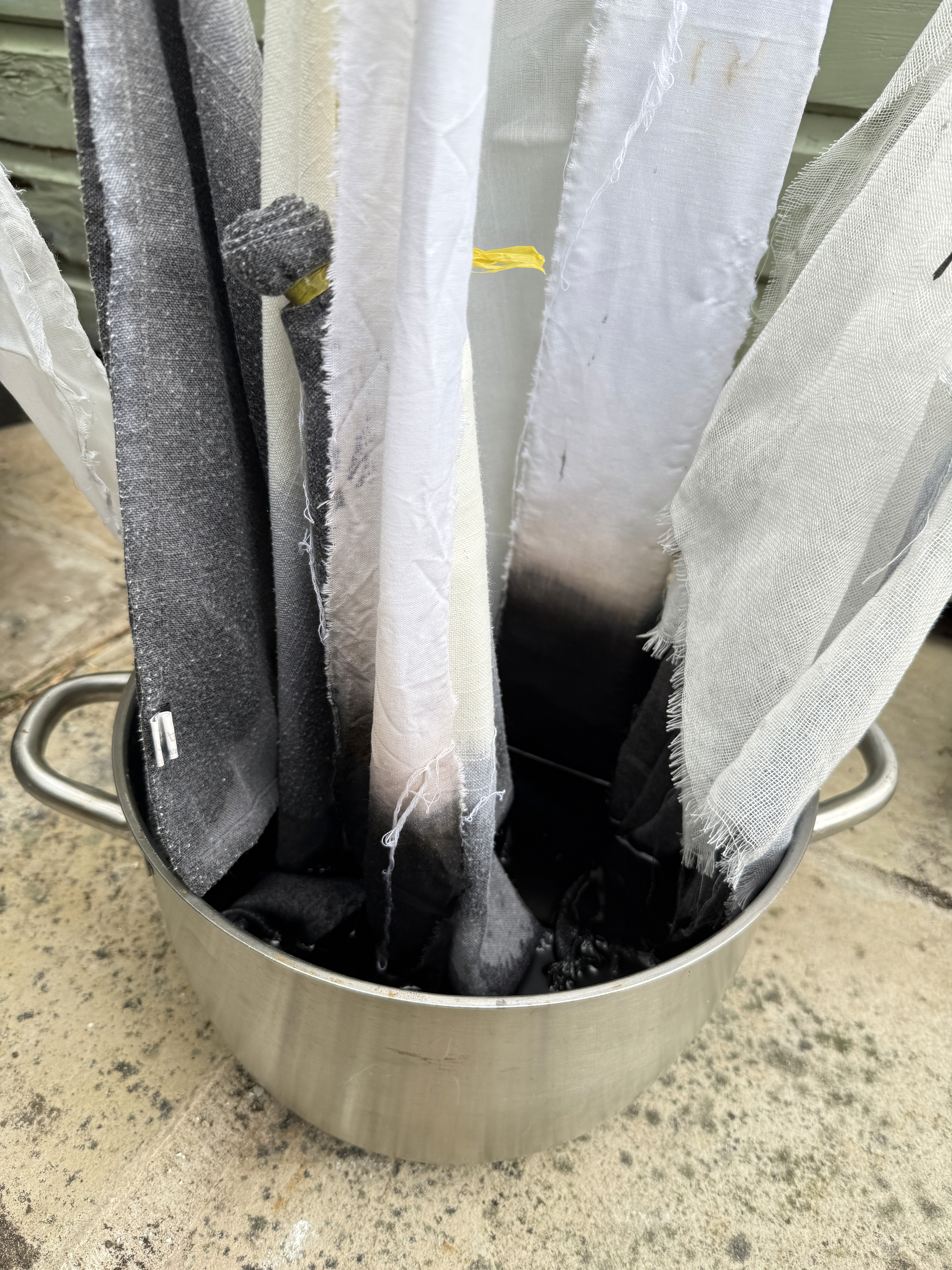 Strips of white and grey fabric are partially submerged in a metal pot of dark dye, showing the gradient transition of colors as the material absorbs the pigment.