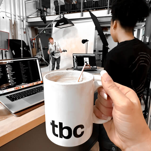 A person holds a white mug labeled tbc in a photo studio, surrounded by laptops, a monitor, and people working in the background—a space buzzing with creative branding and design energy.
