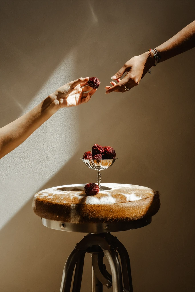 Two hands reaching toward each other above a small table with a dish of red dates