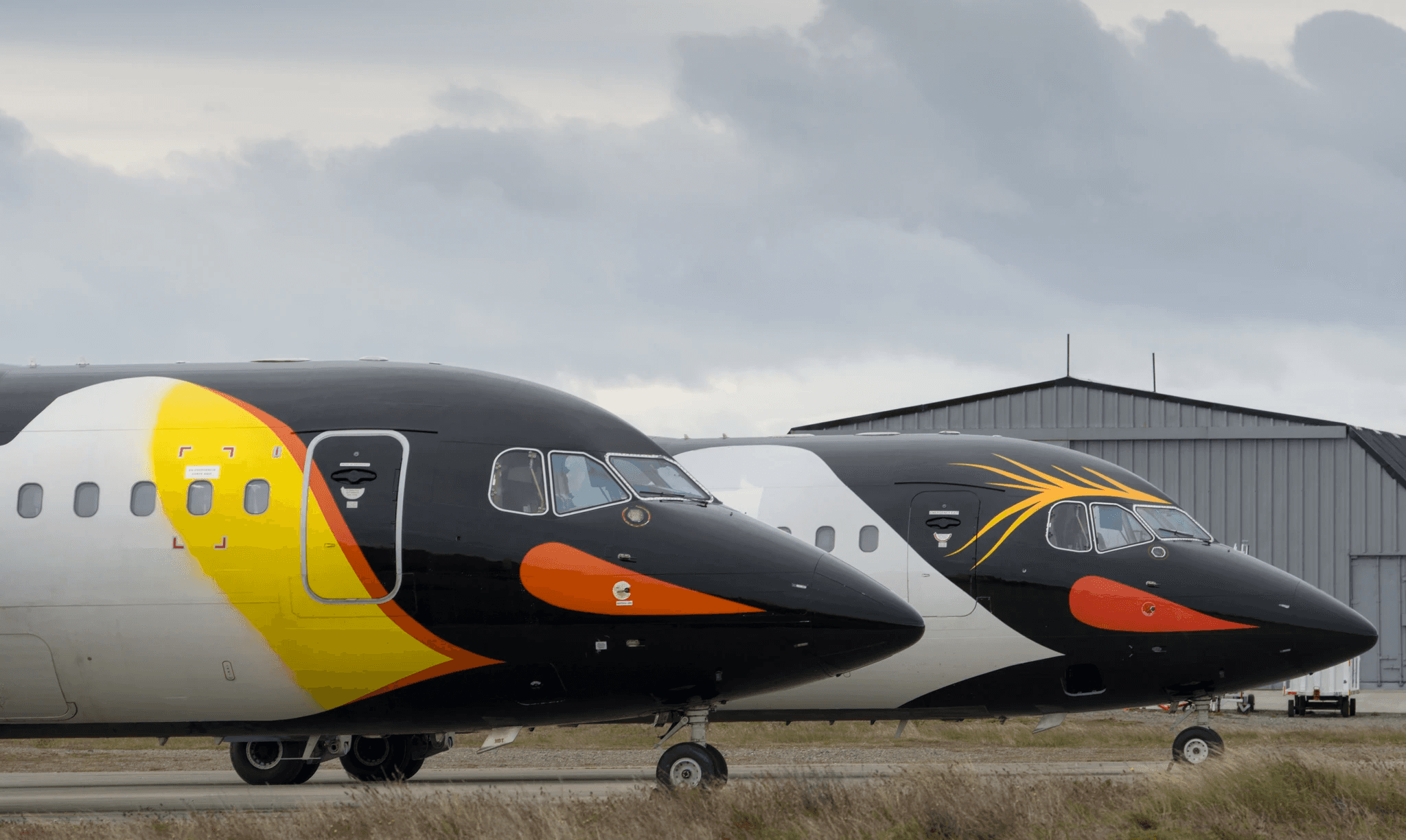 The photograph features two airplanes parked on a runway, positioned side by side. The aircraft in the foreground has a striking black, yellow, and orange color schem