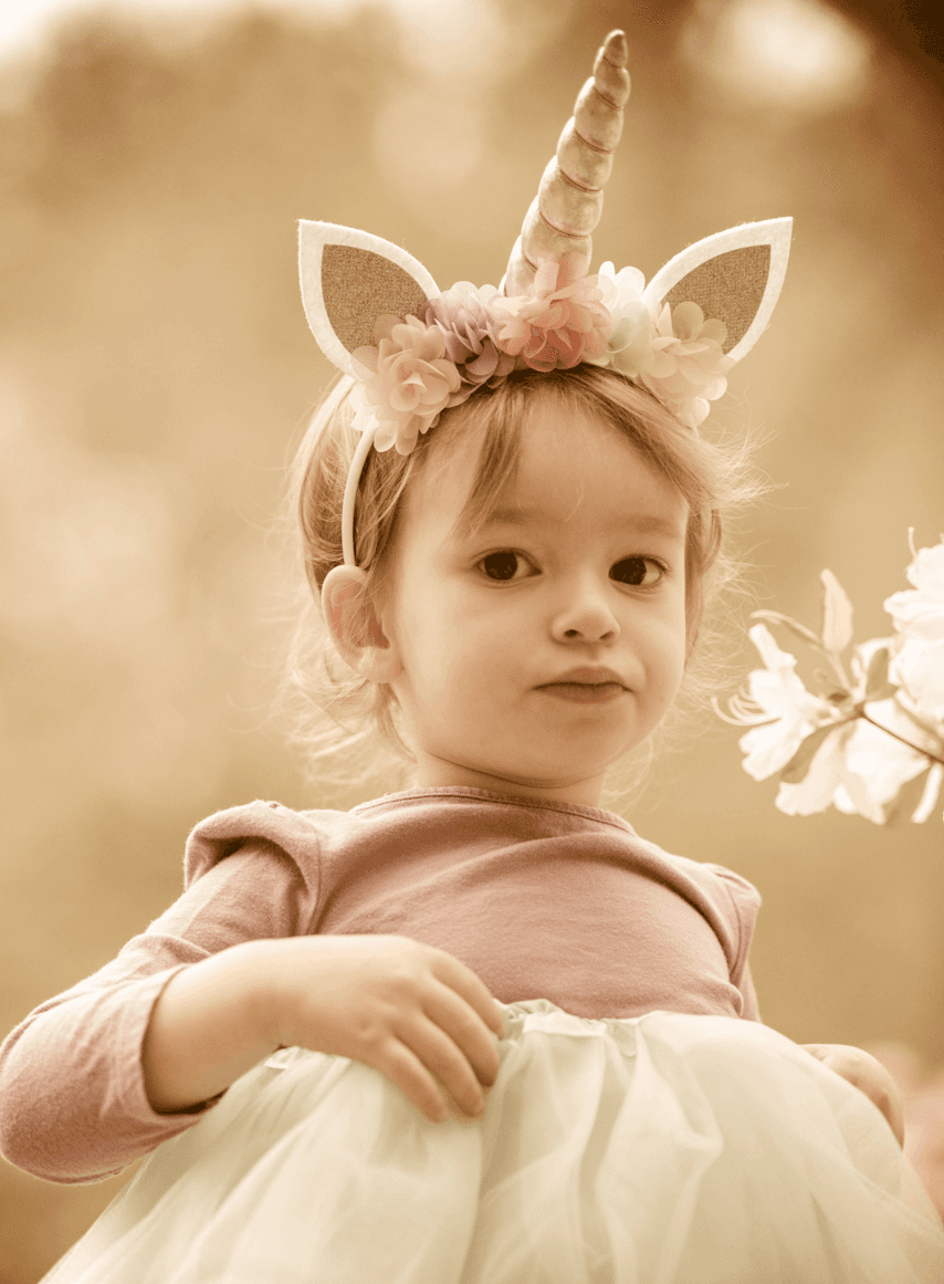 A young child wearing a unicorn headband with flower accents. She's dressed in a pink top and light blue tutu skirt, smiling.