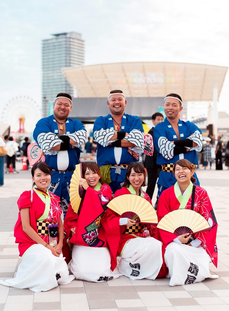 A group of smiling men and women in traditional Japanese festival attire pose together, with the men in blue happi coats and the women in red kimono-style outfits holding fans, against a backdrop of an amusement park and urban structures.