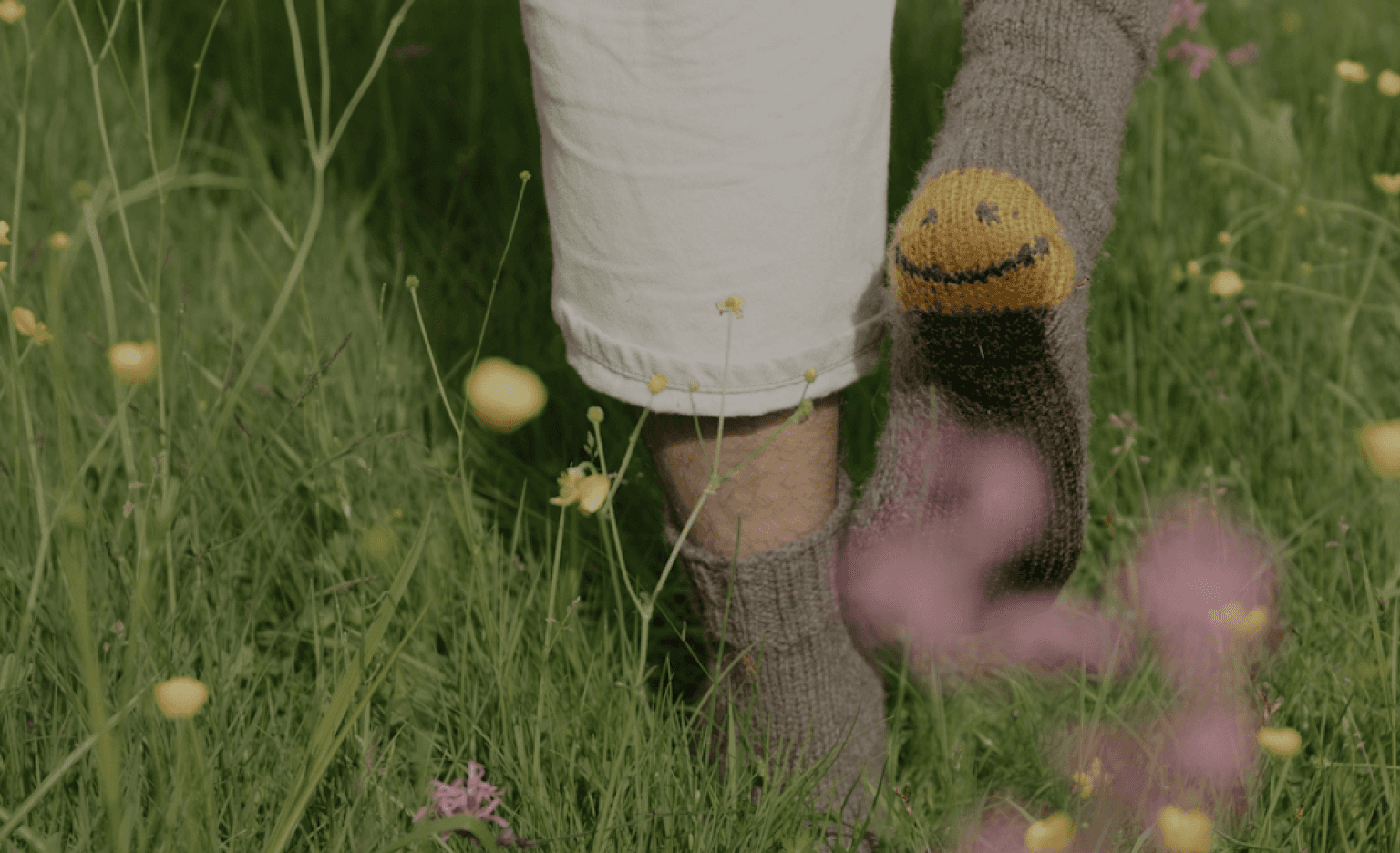 A person's lower body and feet standing in a grassy field filled with yellow and purple flowers, with a knitted sock or leg warmer featuring a smiley face design, creating a serene and whimsical visual composition.