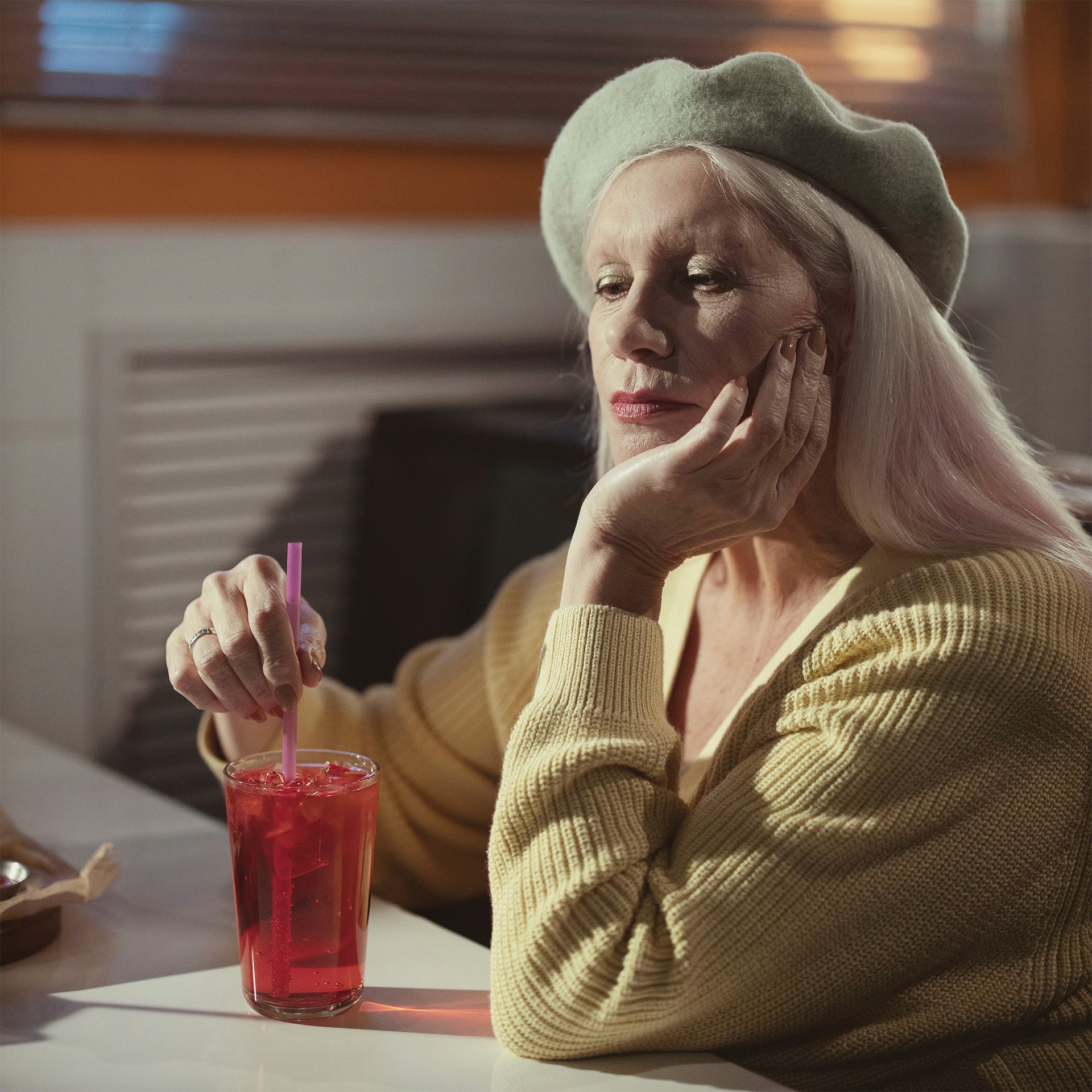 an older woman with blond hair wearing a felt beret sitting in a cafe idly stirring her drink with a straw, her head rests in her other hand and she is lost in thought, slightly sad