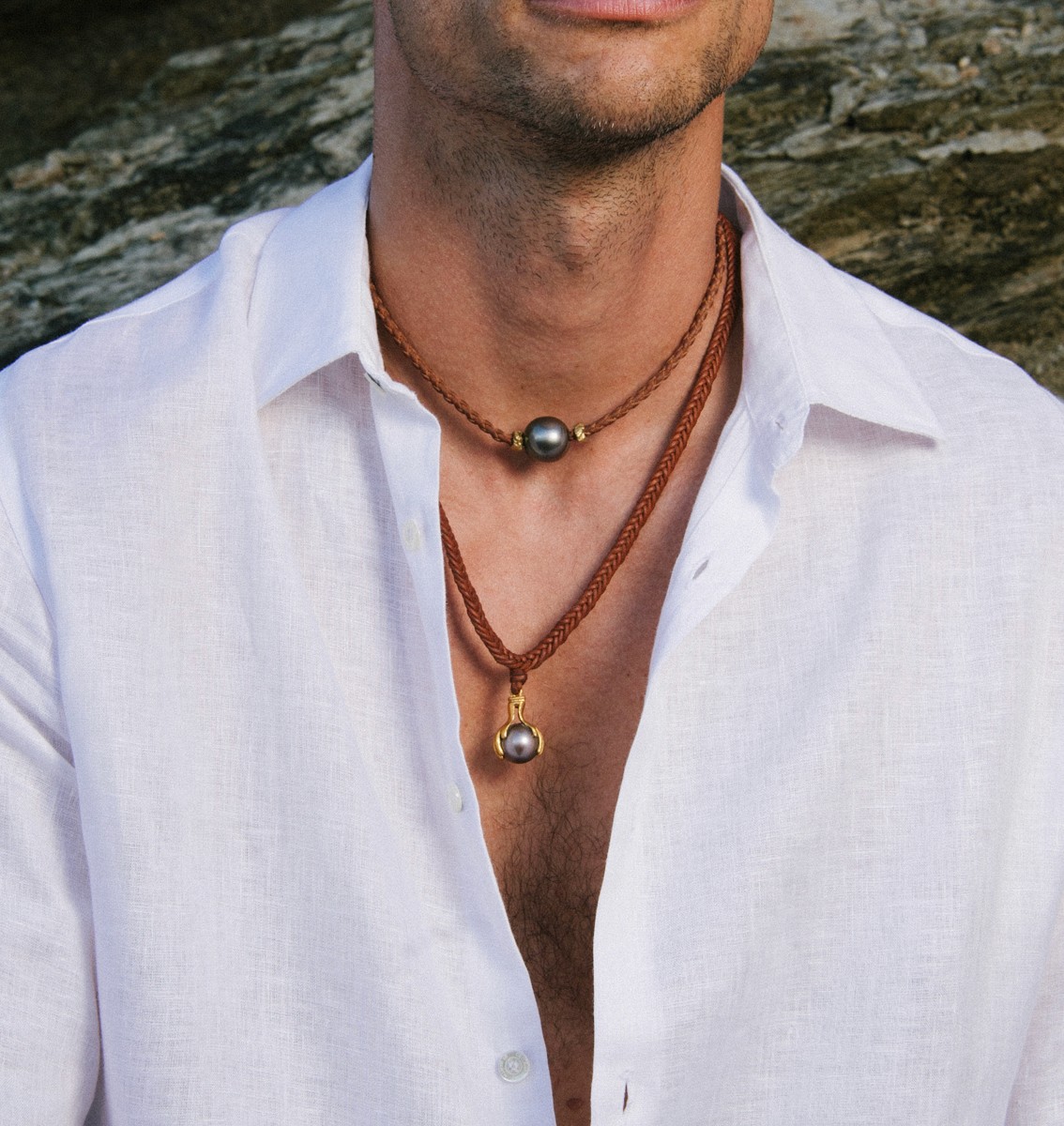 Stylish man wearing layered Tahitian pearl necklaces on braided leather cords, paired with an open white linen shirt for an elegant coastal look.