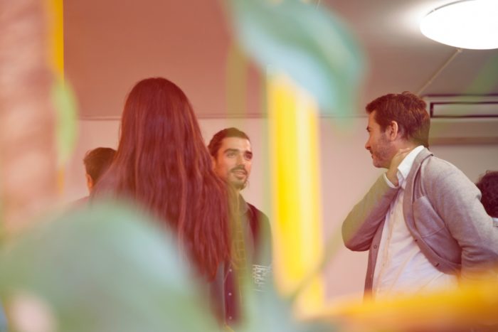 Group of people engaged in conversation indoors, partially obscured by plants and colorful reflections in the foreground.