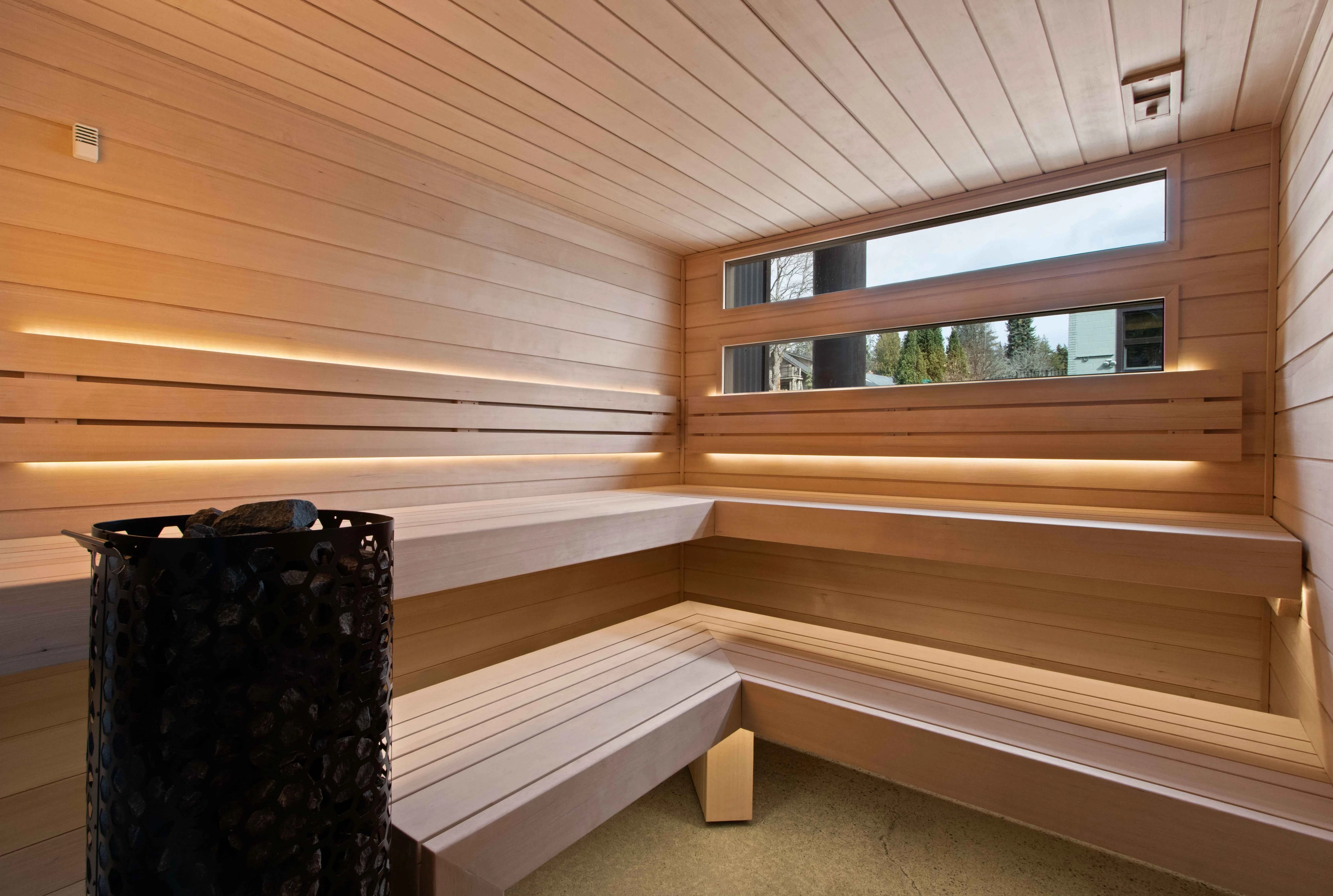Light wood sauna with tiered corner benches, a black stone-filled heater and narrow horizontal windows.
