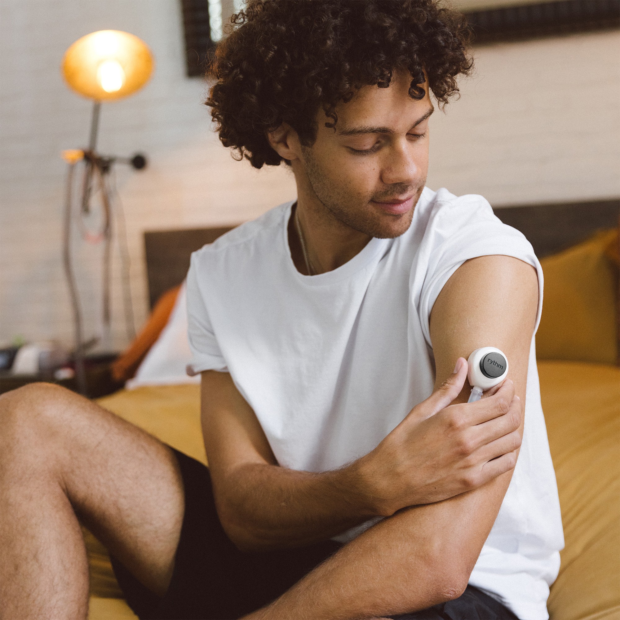 Woman using a Rythm at-home blood collection device on her upper arm, seated indoors in soft natural light, looking down as she applies the device.