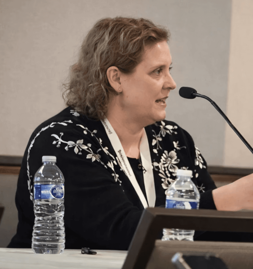A smiling man in a dark shirt and jeans sits casually on a stool against a dark grey background, looking off to the side.