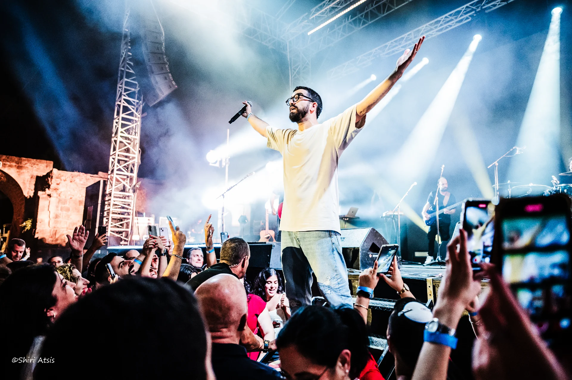 צילום מרגש מתוך הופעה חיה של חנן בן ארי, האמן עומד על קצה הבמה בידיים פרושות מול קהל מעריצים שמצלם ושר איתו יחד באווירה מחשמלת, מאת Shiriz Photography הצלמת שירי עציץ.