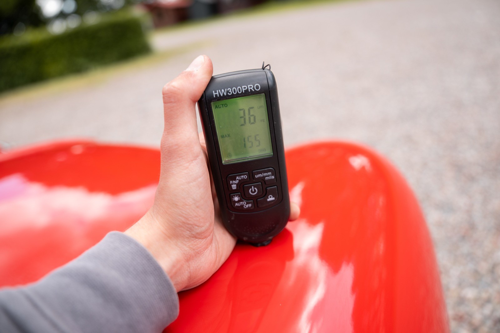 A hand holds a device displaying data on a red surface, with a blurred background of greenery.