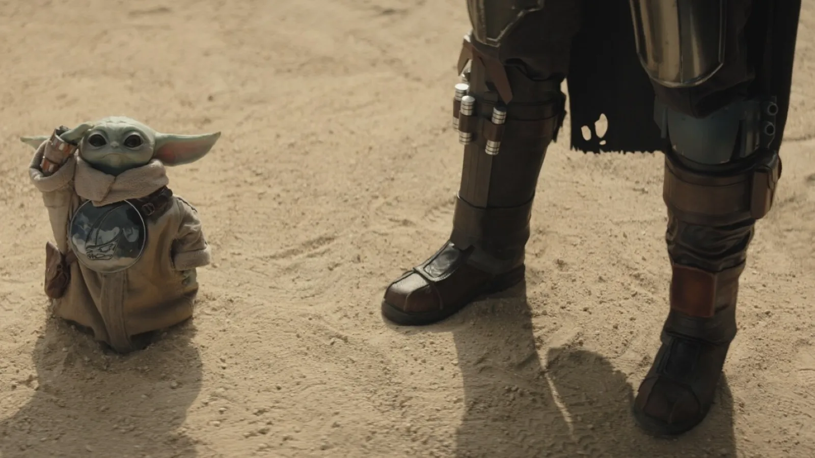 A small green alien creature stands on sandy terrain, cloaked in a beige robe, next to a person wearing metallic armour, showcasing a scene of adventure and exploration in a desert setting.
