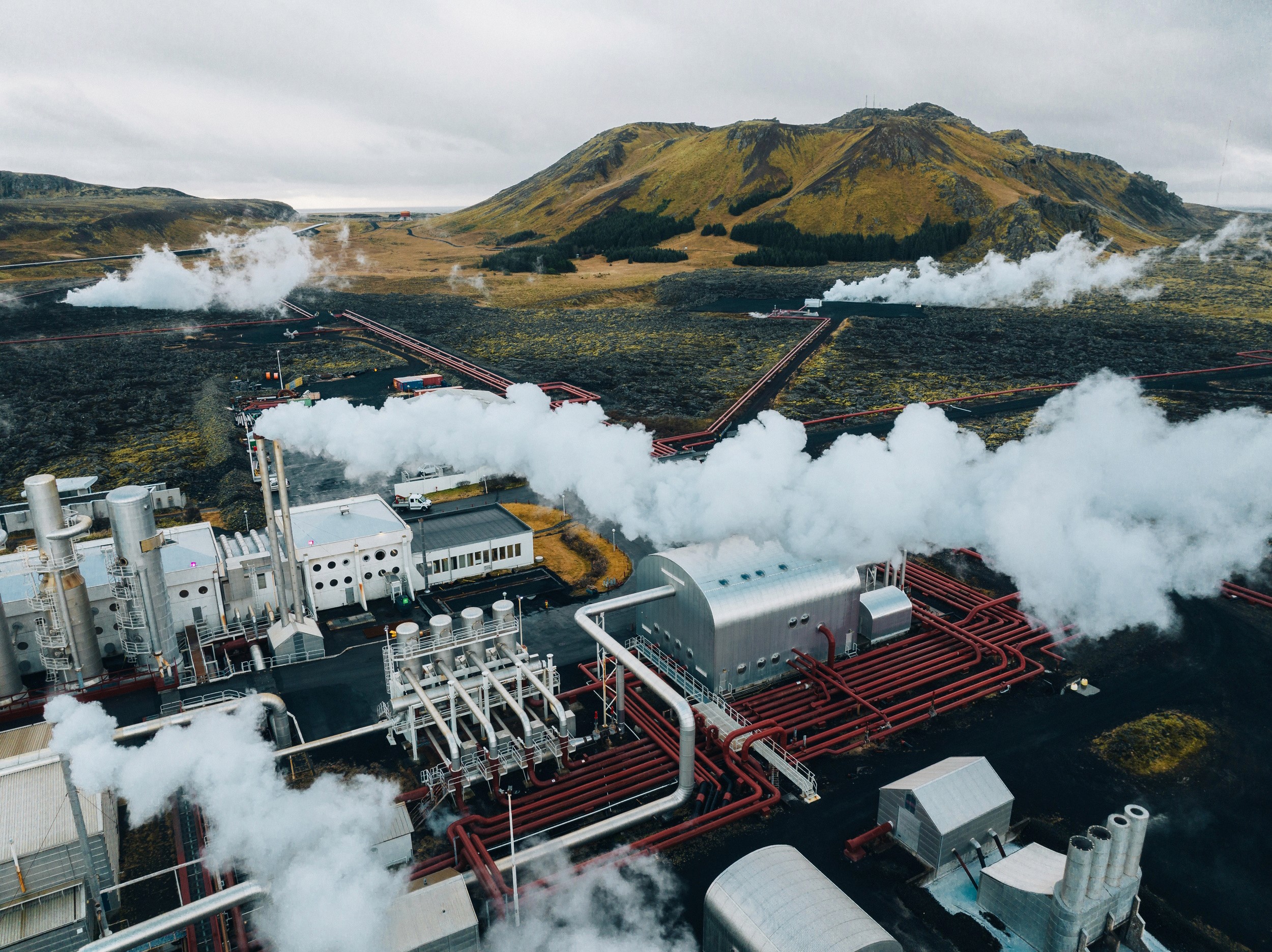 Aerial view of the HS Orka geothermal power plant near Grindavík, Iceland, with steam rising and red geothermal pipelines distributing hot water across the black lava landscape.
