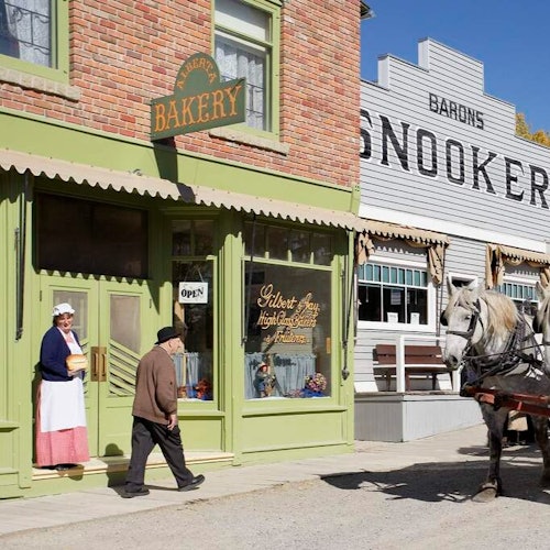 A woman in period costume stands outside a bakery while a man walks toward it; a horse-drawn carriage is nearby.