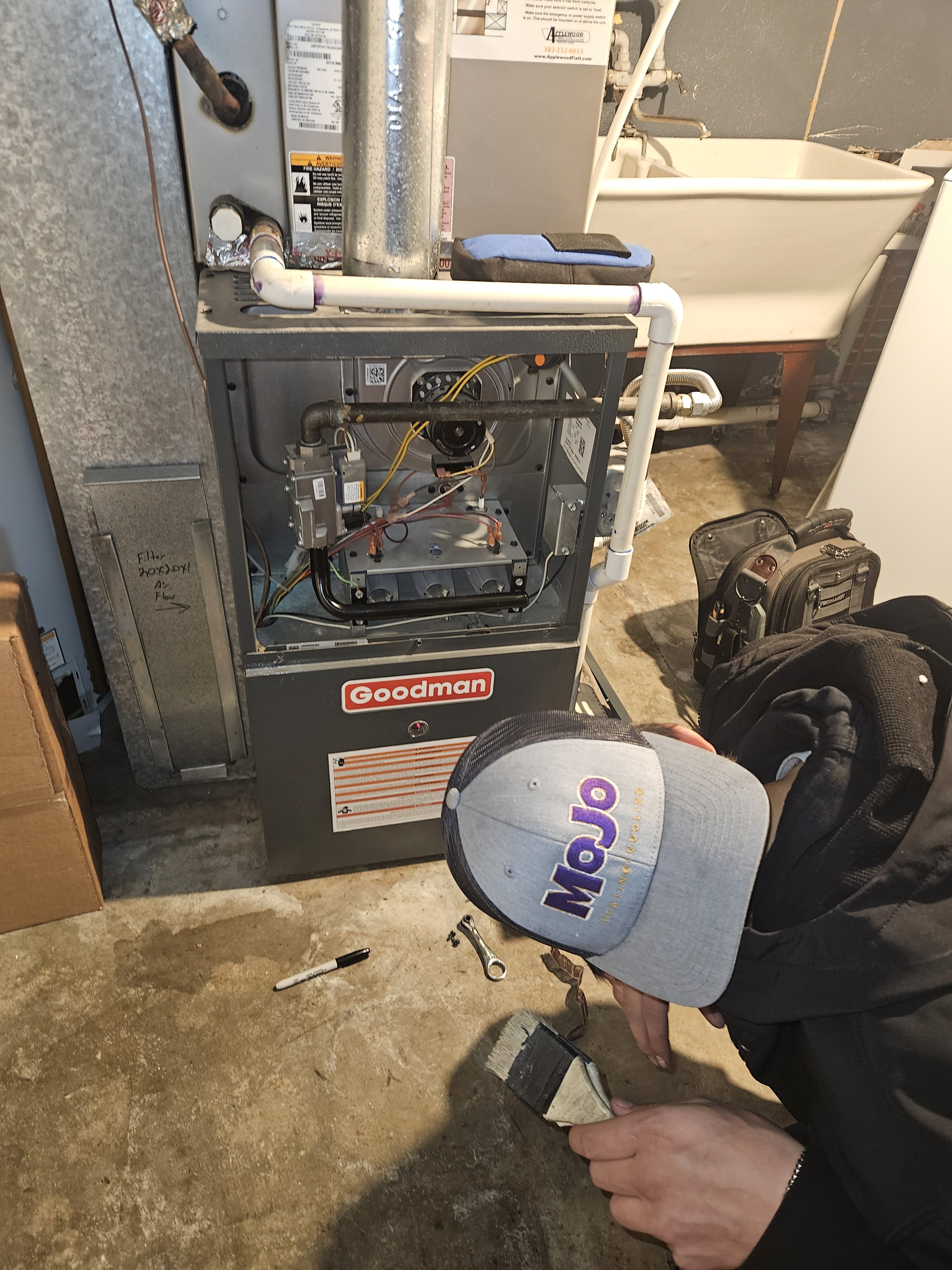 The image shows MOJO HVAC technician servicing a Goodman gas furnace. The technician is likely employed by MoJo Home Services, a company that provides heating installation and maintenance services in the Denver, CO area
