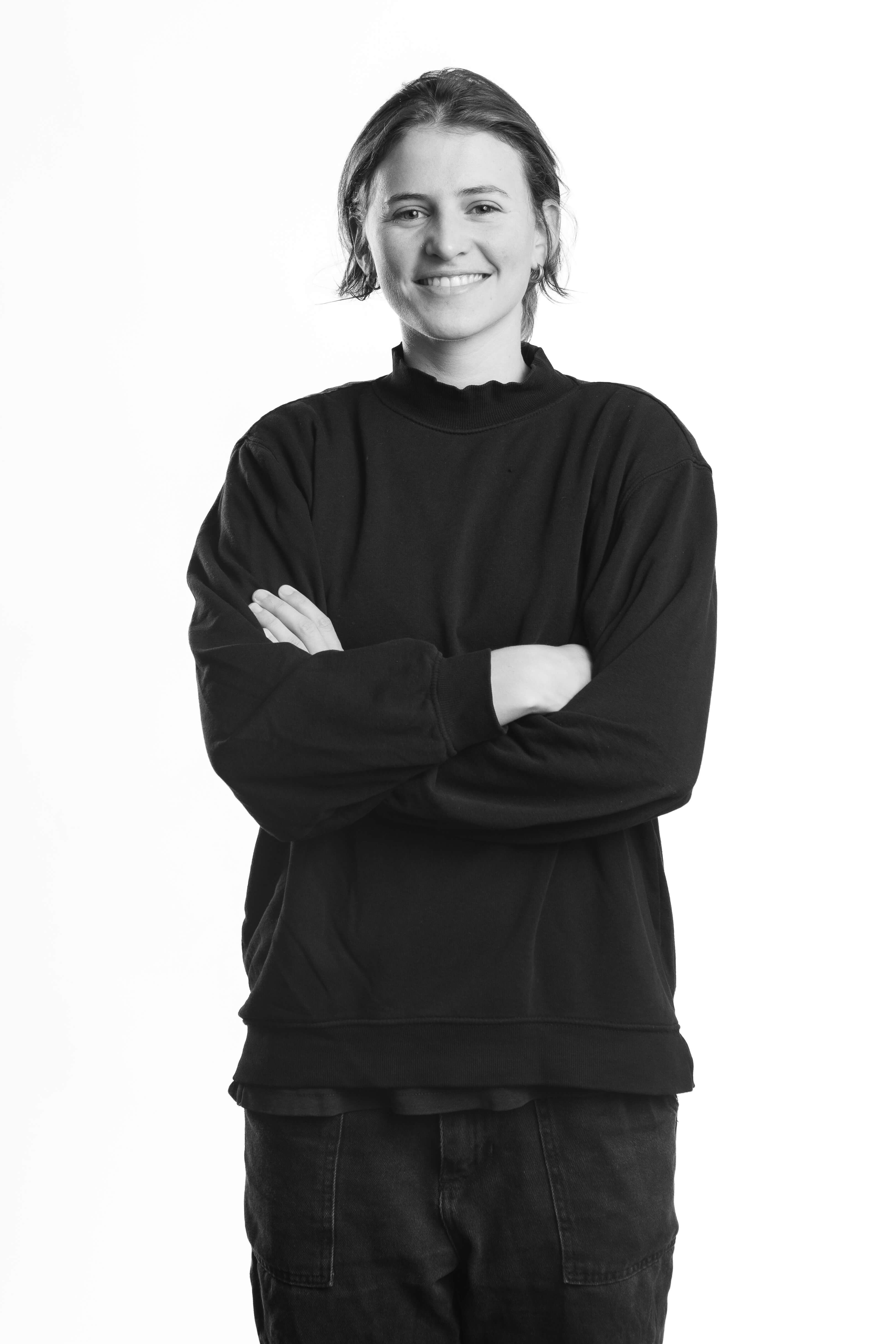 Black and white image of a person with short hair, smiling and standing with arms crossed. They wear a dark sweater and jeans, conveying a relaxed, confident tone.