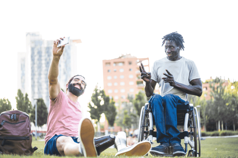 Two friends sitting on grass outdoors, taking a selfie; one is using a wheelchair.