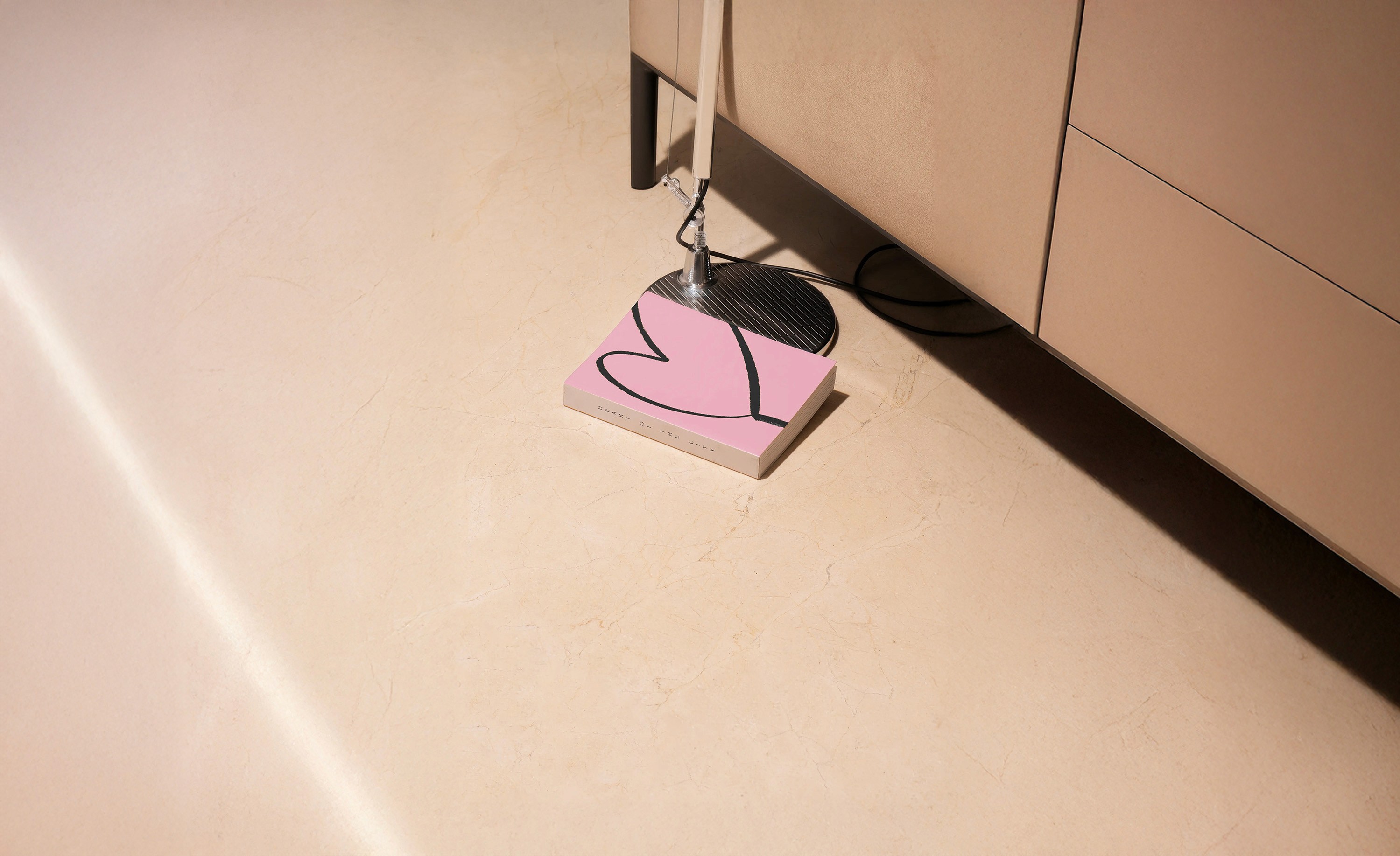 A soft pink book titled Heart of the City rests at the base of a chrome floor lamp, grounding its delicate cover beneath the shadows of a modern interior. A bold heart line dances across the surface — simple, intuitive, human. A quiet presence in an urban space, evoking intimacy, longing, and the pulse of love amid concrete stillness. From Zapersona’s Creative Archive — a visual love letter to inner-city emotion and expressive minimalism. Creative Expression, Visual Poetry.