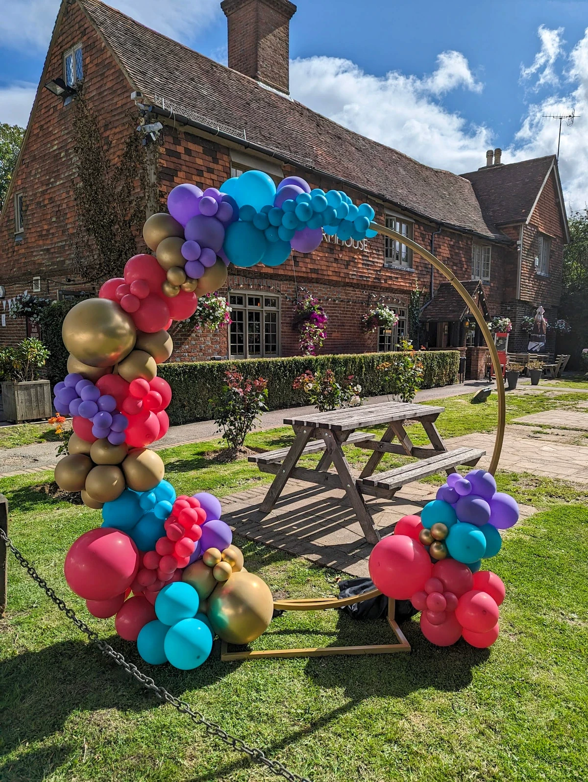 Outdoor garden balloon arch with a mix of red, teal, purple, and gold balloons