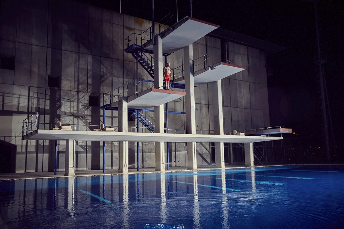 Lone diver in red shorts standing on high platform diving board above glowing blue Olympic swimming pool at night, dramatic brutalist aquatic centre architecture