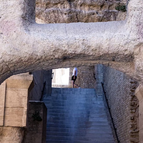 Một người mặc quần short đi lên cầu thang đá dài dưới một cổng vòm đá lớn, với những bức tường gạch cổ xưa ở cả hai bên.