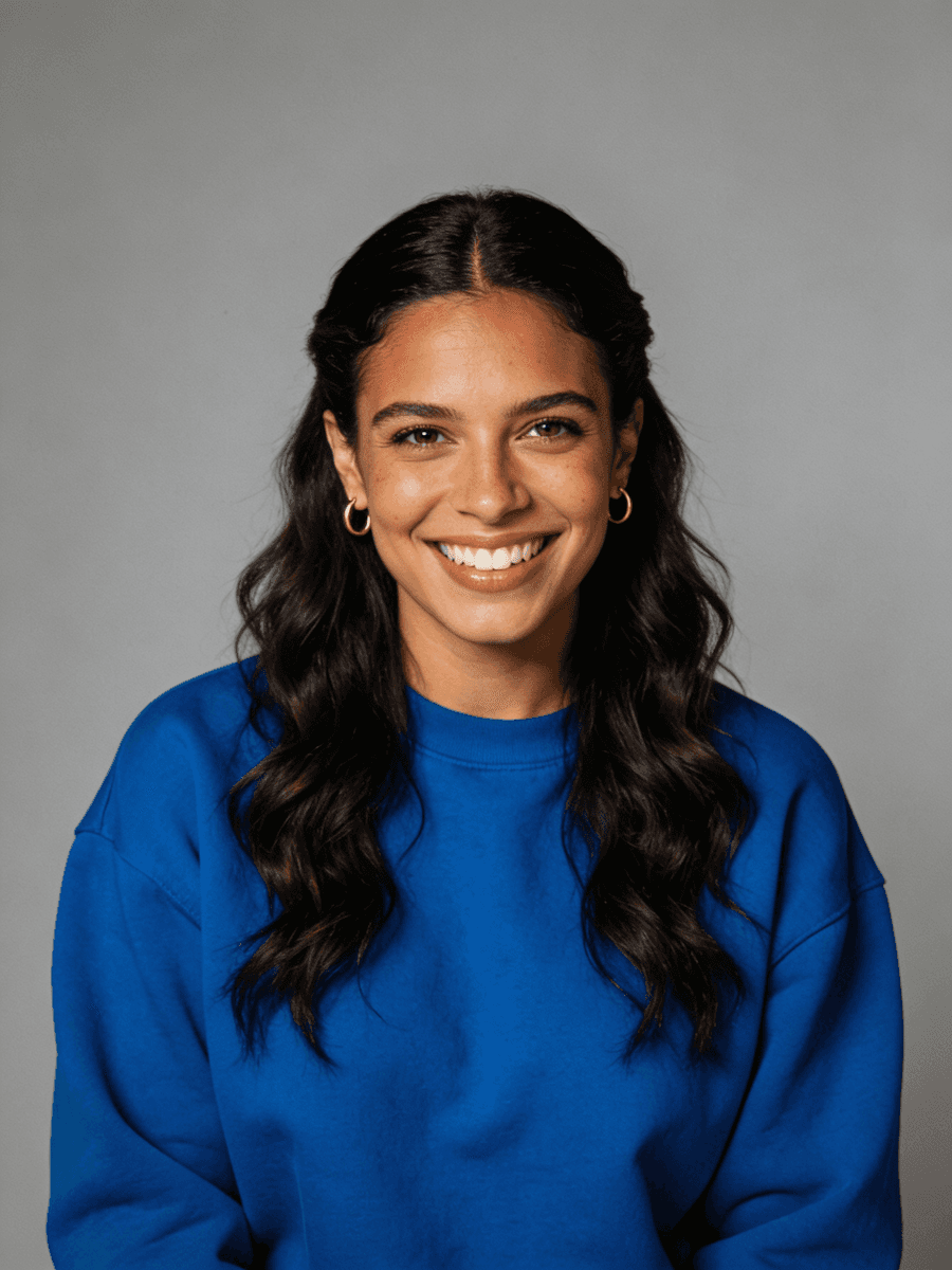 Portrait of a smiling woman with long wavy hair wearing a blue sweatshirt and gold hoop earrings against a gray background.
