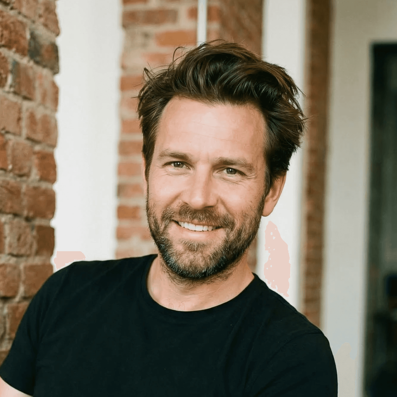 Smiling man with brown hair and a beard wearing a black t-shirt, standing in front of a brick wall.