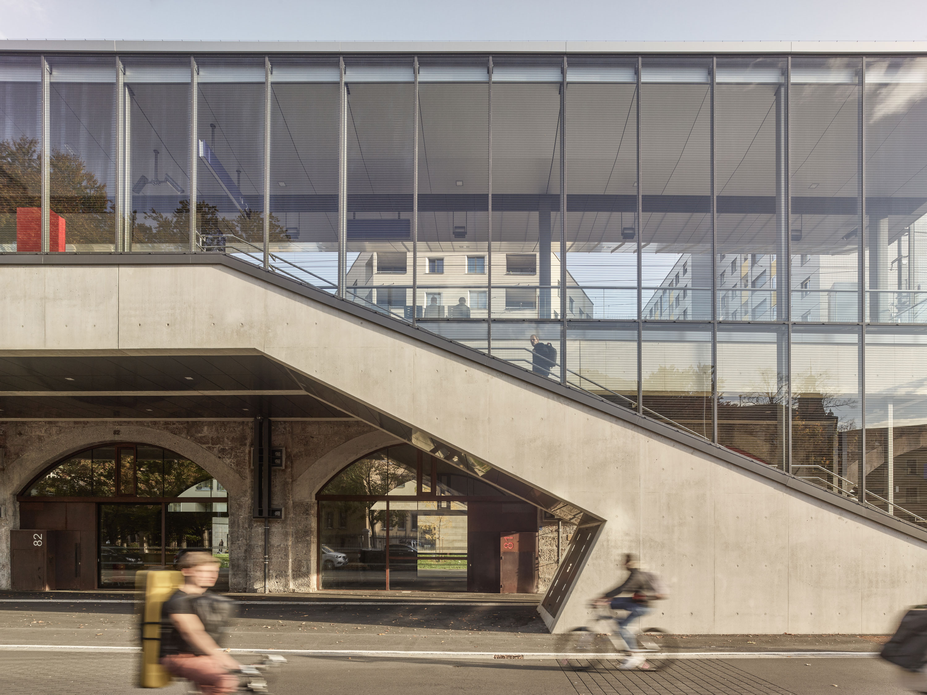 Moderner Bahnhof Innsbruck Messe mit gläserner Fassade und Sichtbetontrampe, zwei Personen auf Fahrrad im Vordergrund.