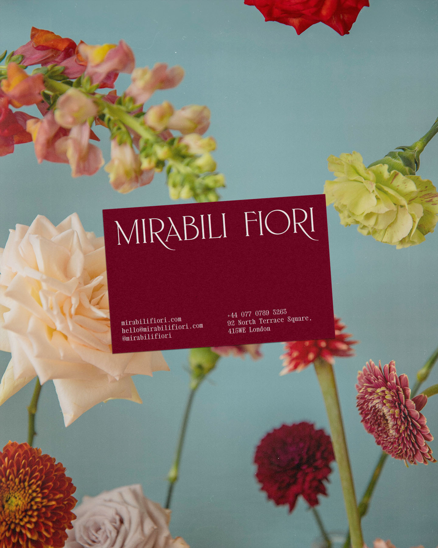Burgundy business card among fresh roses, dahlias, and carnations on blue background