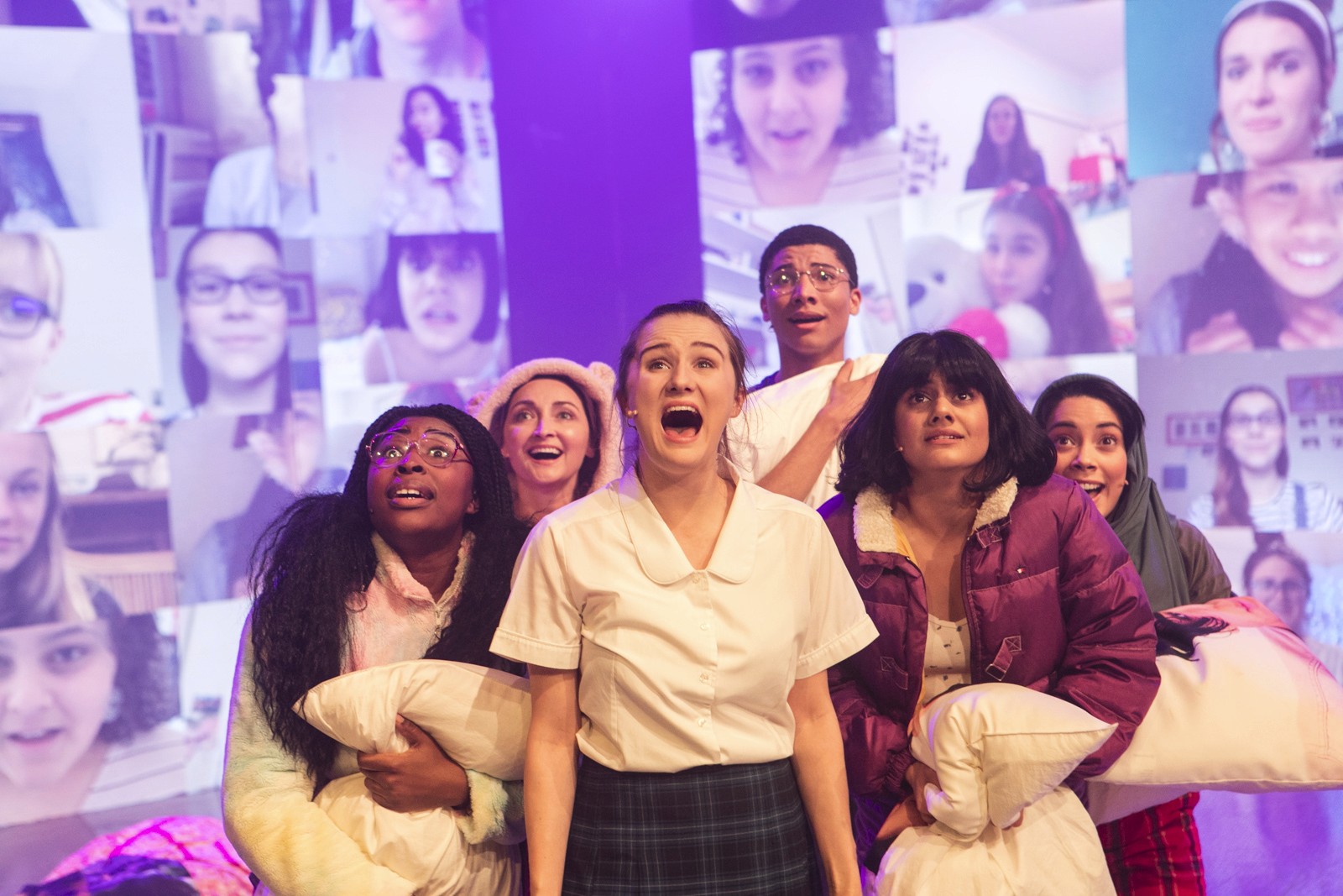 Six young performers clutch pillows and blankets, looking upwards in awe, as a digital wall of fan webcam videos glows behind them on stage.