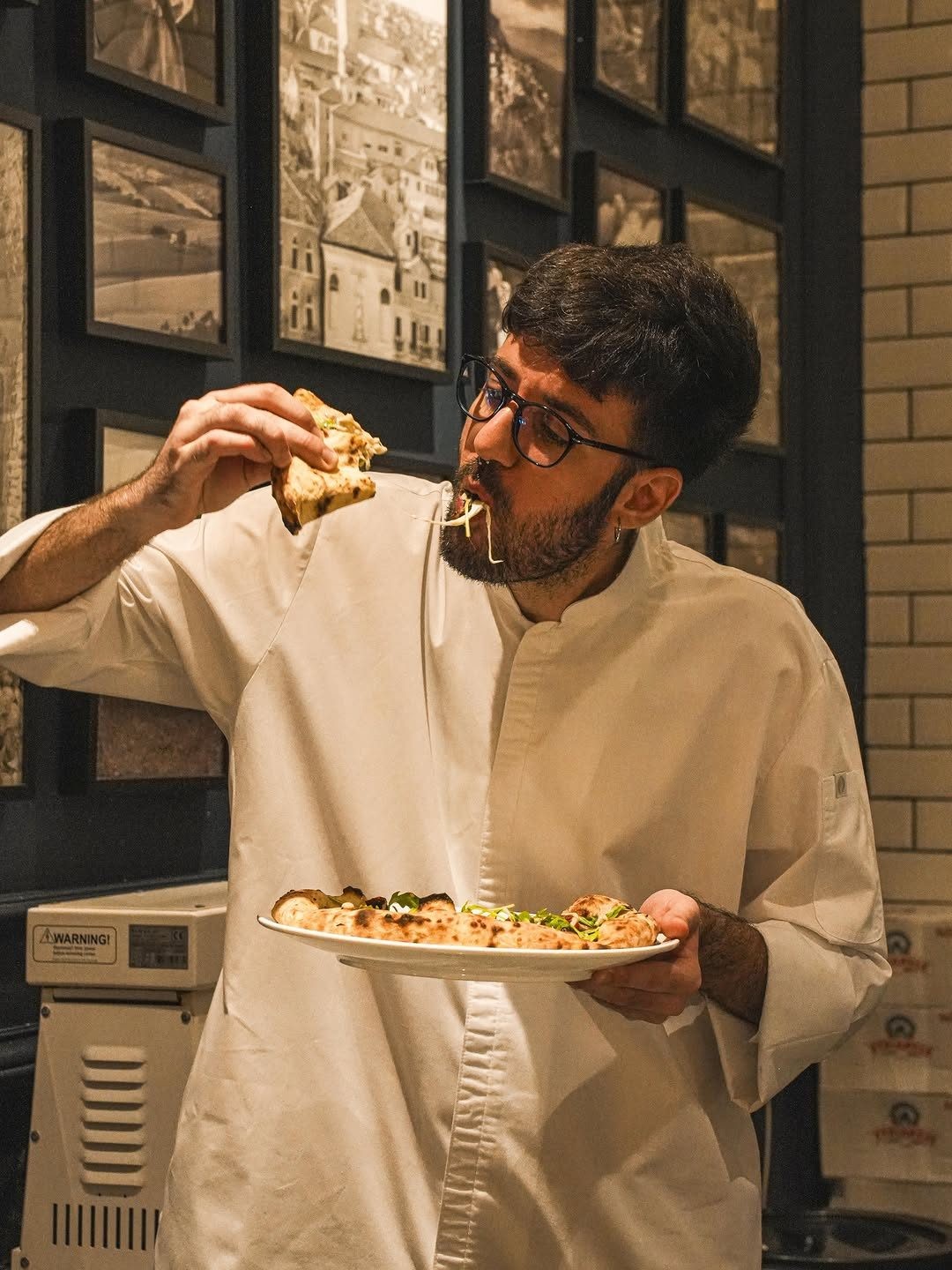 A chef in a white coat holds a plate of food while taking a bite from a piece of pizza, with framed black and white photographs in the background.