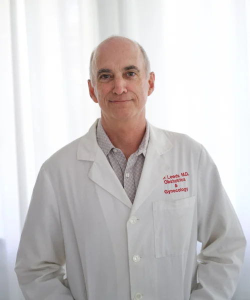 Doctor in a white medical coat with embroidered text for obstetrics and gynecology, shown in front of light curtains in a clinical environment.