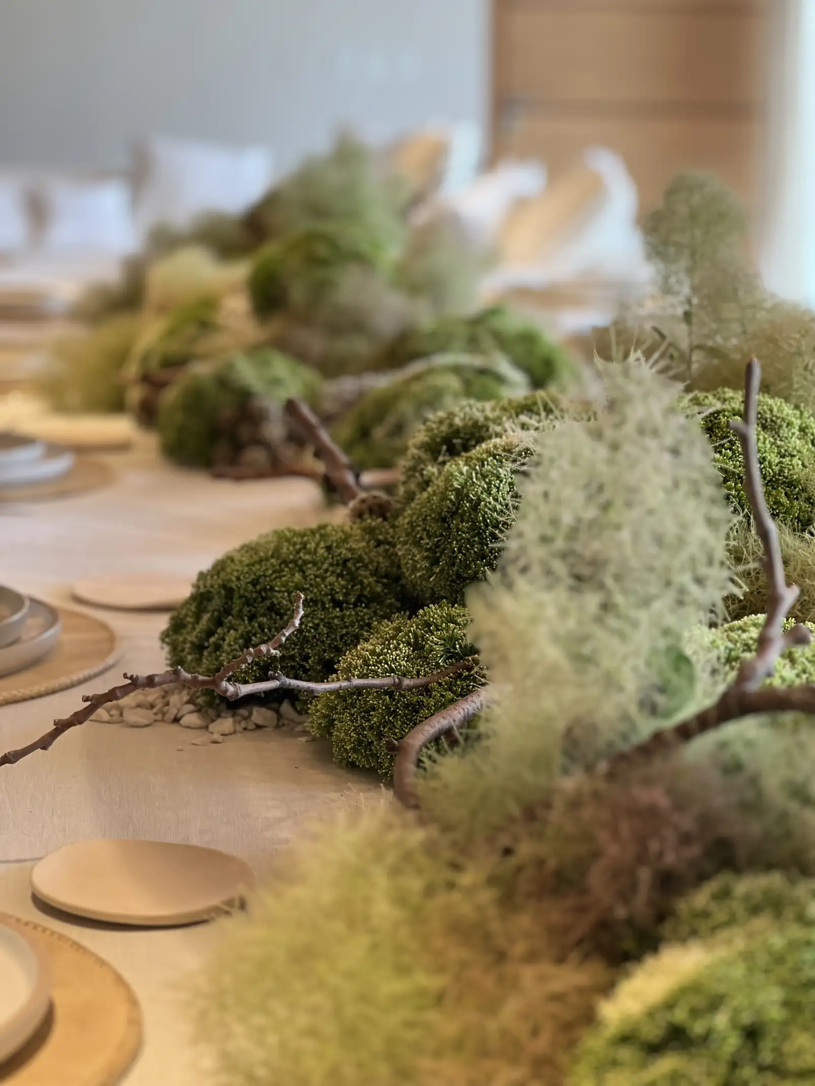 Vista panorámica de mesa larga con decoración de musgos y ramas, cristalería delicada y lámparas de mimbre colgantes.