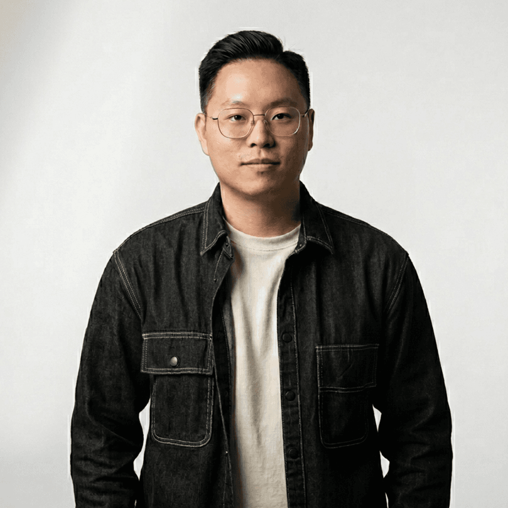 A medium-shot portrait of an Asian man with short, black, styled hair, wearing thin silver-rimmed glasses, a dark charcoal denim button-down jacket over a cream t-shirt, looking directly at the camera with a neutral expression, set against a plain light grey studio background.
