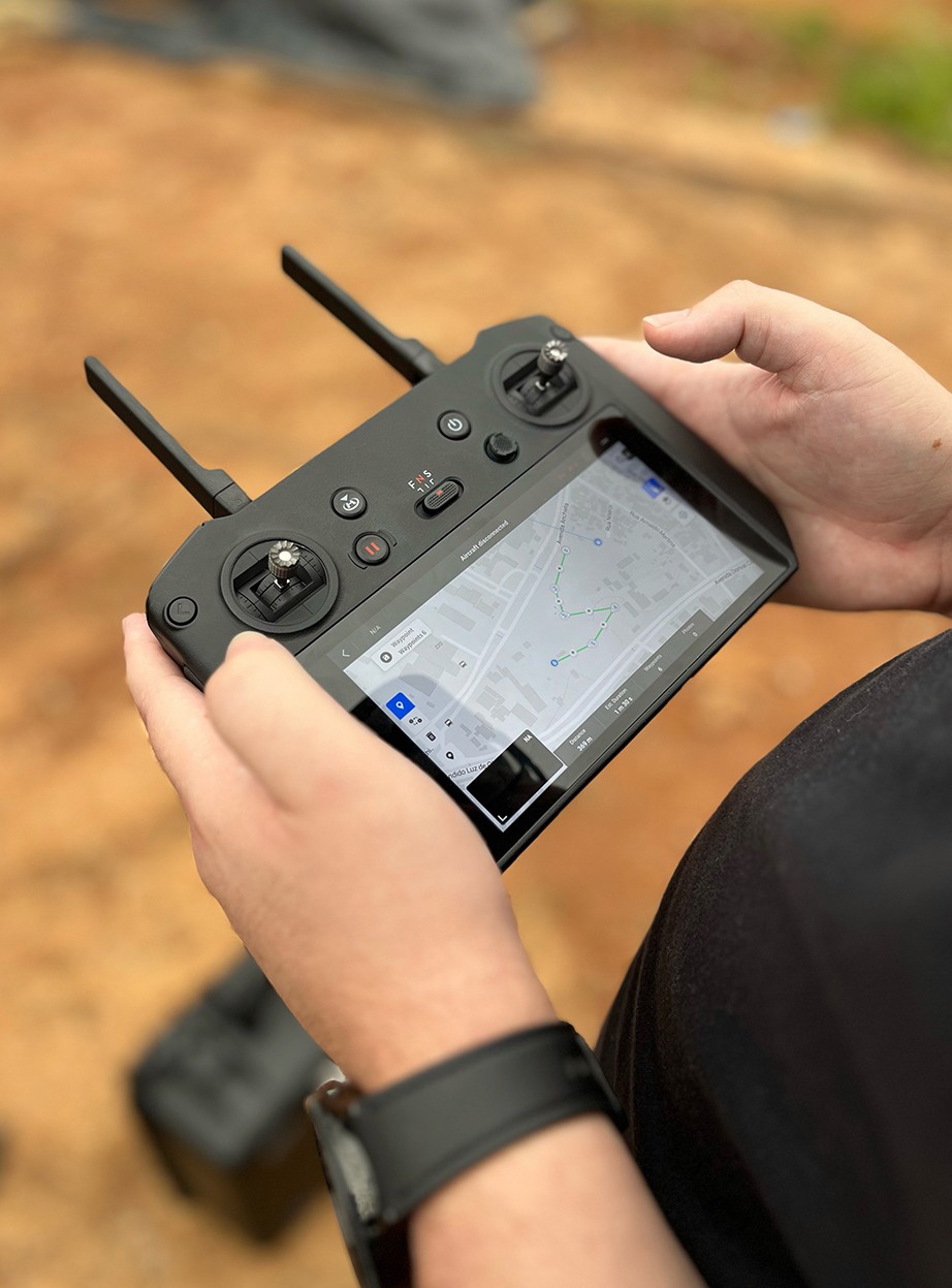 Mãos operando um controle remoto de drone que exibe um mapa com uma rota de voo planejada por pontos (waypoints). O recurso demonstra a automação da Captura da Realidade para garantir que as fotos aéreas sejam tiradas sempre dos mesmos ângulos.