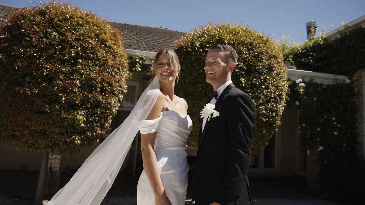 A smiling bride in an elegant white strapless gown with a flowing veil stands next to a groom in a classic black tuxedo, surrounded by lush green foliage under a clear blue sky, showcasing a joyous wedding scene.