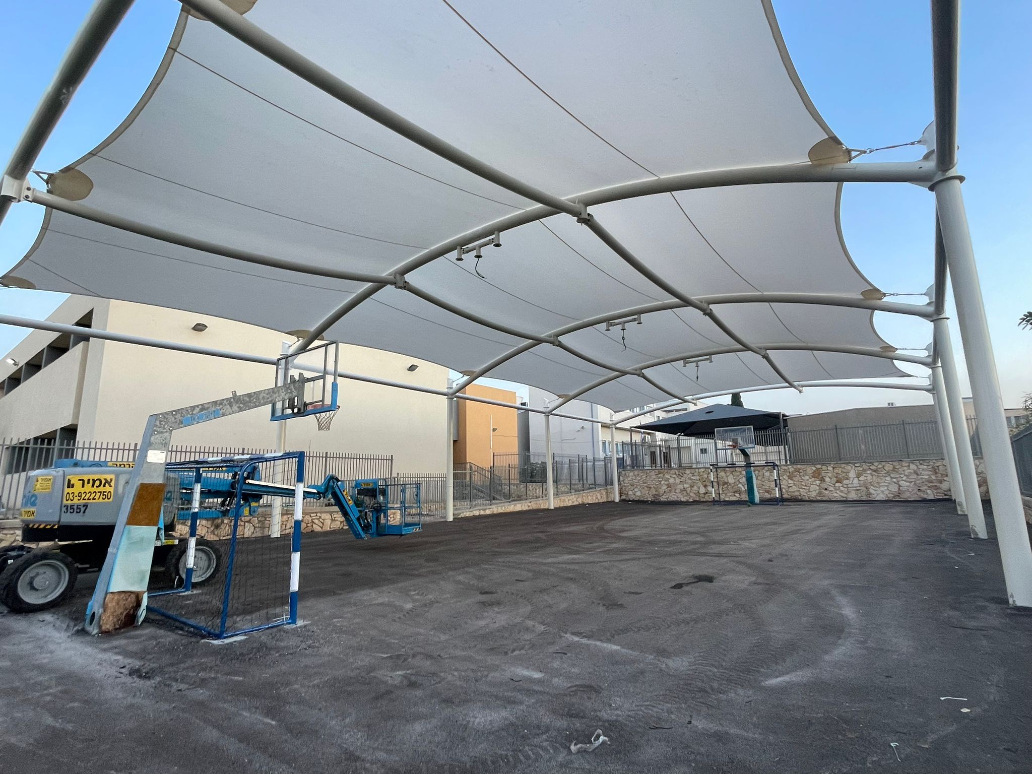 Covered outdoor area with two basketball hoops and a construction vehicle on asphalt near a fence and buildings.