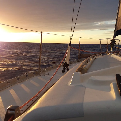 Solnedgångsvy från däck på en segelbåt på lugnt vatten, med rigg och segel synliga.