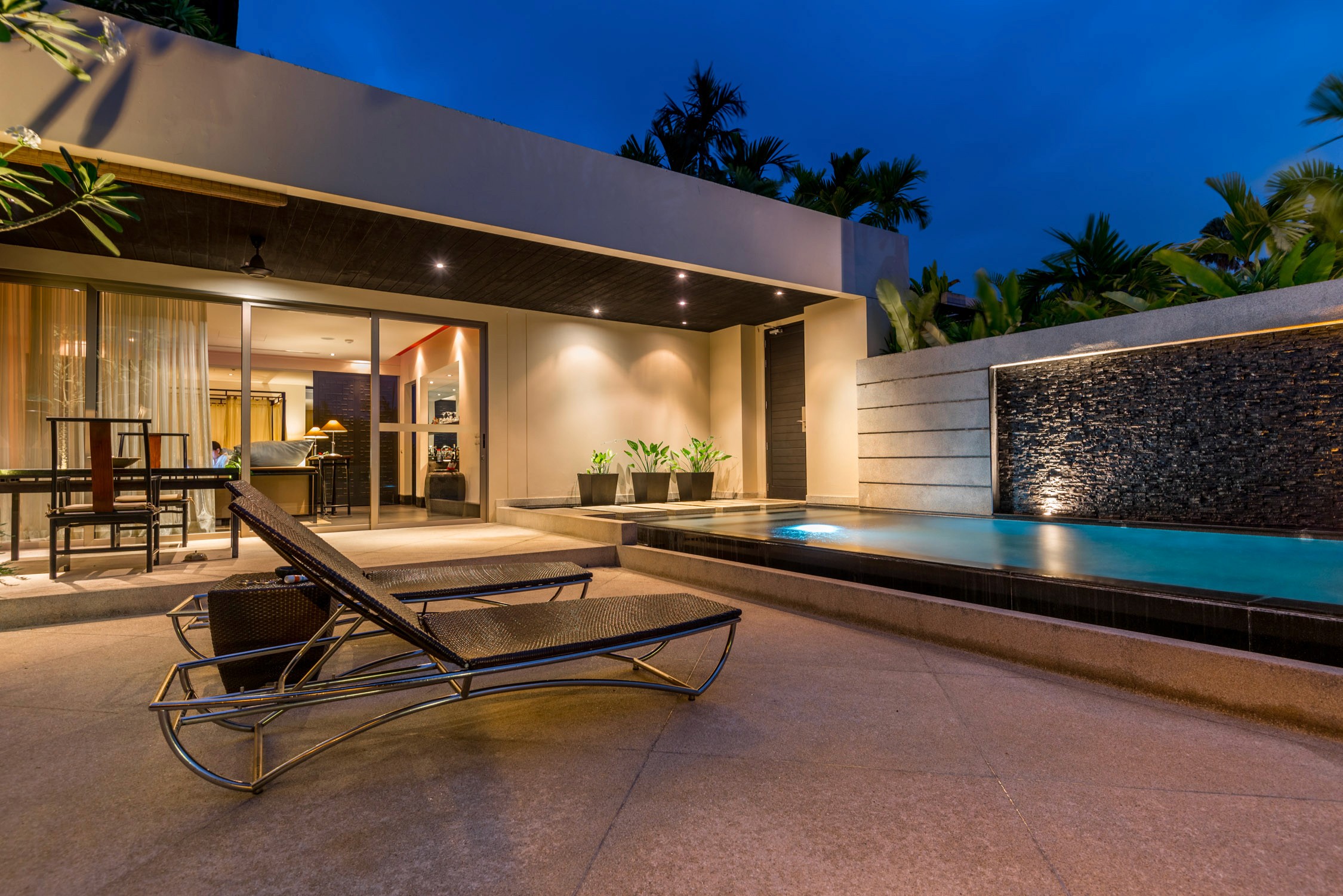 A modern poolside lounge at dusk features a lit swimming pool, two reclining chairs, and an open-air dining area.